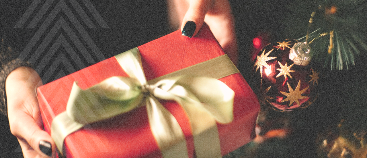 Person holding a wrapped holiday gift beside a decorated Christmas tree with a red and gold ornament.
